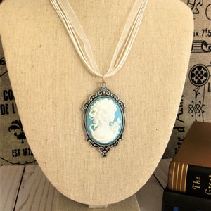May include: A cameo pendant necklace with a white profile on a light blue background, set in a silver-toned frame. The pendant hangs from a white ribbon necklace. The necklace is displayed on a beige mannequin.