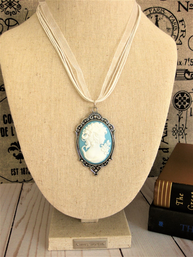 May include: A cameo pendant necklace with a white profile on a light blue background, set in a silver-toned frame. The pendant hangs from a white ribbon necklace. The necklace is displayed on a beige mannequin.