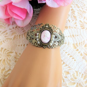 May include: A delicate gold-tone bracelet featuring a cameo with a pink profile of a woman. The bracelet is decorated with faux pearls and intricate filigree details.