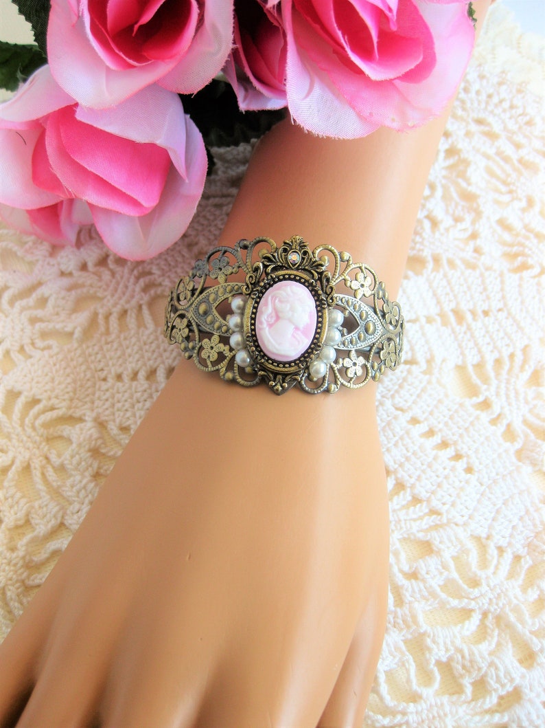 May include: A delicate gold-tone bracelet featuring a cameo with a pink profile of a woman. The bracelet is decorated with faux pearls and intricate filigree details.