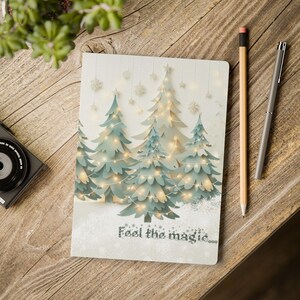 Puede incluir: Una libreta blanca con una escena de invierno de árboles verdes con luces blancas y copos de nieve. El texto "Feel the magic..." está impreso en la parte inferior de la libreta.
