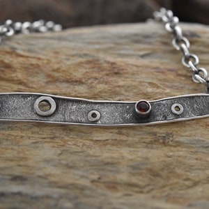 May include: A silver necklace with a textured, elongated pendant featuring a red gemstone and several circular cutouts. The pendant is attached to a silver chain.
