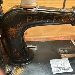 Peut inclure: Une machine à coudre vintage noire avec des accents dorés et le mot "PFAFF" sur le bras. La machine a un motif floral décoratif sur le côté.