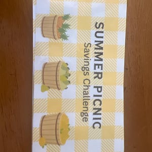 May include: A rectangular card with a yellow and white checked pattern. The card features three illustrations of wooden baskets filled with fruit. Text on the card reads "SUMMER PICNIC Savings Challenge."