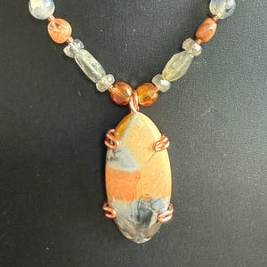 May include: A handcrafted necklace featuring a large, oval pendant with orange, gray, and tan hues, secured by copper wire. The necklace includes a beaded chain with various light-colored stones and orange beads, set against a dark background.