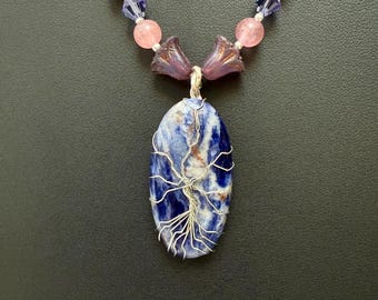 May include: A silver wire wrapped tree of life pendant with a blue and white sodalite stone. The pendant is attached to a necklace with pink and purple beads.