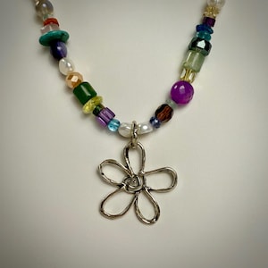 May include: A colorful beaded necklace with a silver flower pendant. The necklace features a variety of beads in different colors, shapes, and sizes, including turquoise, amethyst, and green jade. The flower pendant is delicate and intricate, with a swirl design in the center.