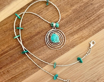 May include: A silver necklace with a turquoise pendant. The pendant is a circle with multiple concentric circles around it. The necklace is made of small silver beads and has turquoise stones spaced along the chain.