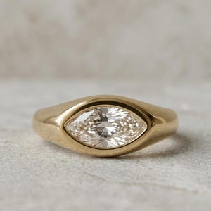 May include: A gold ring featuring a single marquise-cut clear gemstone set horizontally in a smooth, polished bezel setting. The band is solid gold with a rounded profile, tapering slightly toward the back, showcasing a clean and reflective finish.