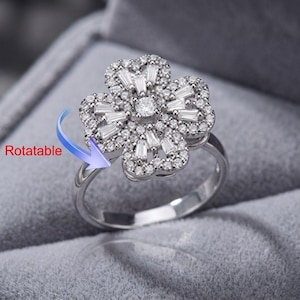 May include: A silver-toned ring featuring a flower-shaped centerpiece adorned with clear baguette and round stones. The floral element is designed to be rotatable, as indicated by a blue arrow graphic and the text Rotatable on the side.