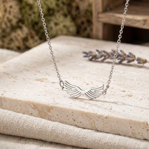May include: A silver-toned necklace featuring a central pendant shaped like a pair of symmetrical angel wings. The pendant is attached to a delicate, thin metal chain with small circular links that extend upward from each side of the wing design.