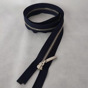 May include: A navy blue zipper with a silver metal pull and teeth. The zipper is coiled and partially unzipped, showing the metal teeth. Suitable for sewing projects and garment construction.