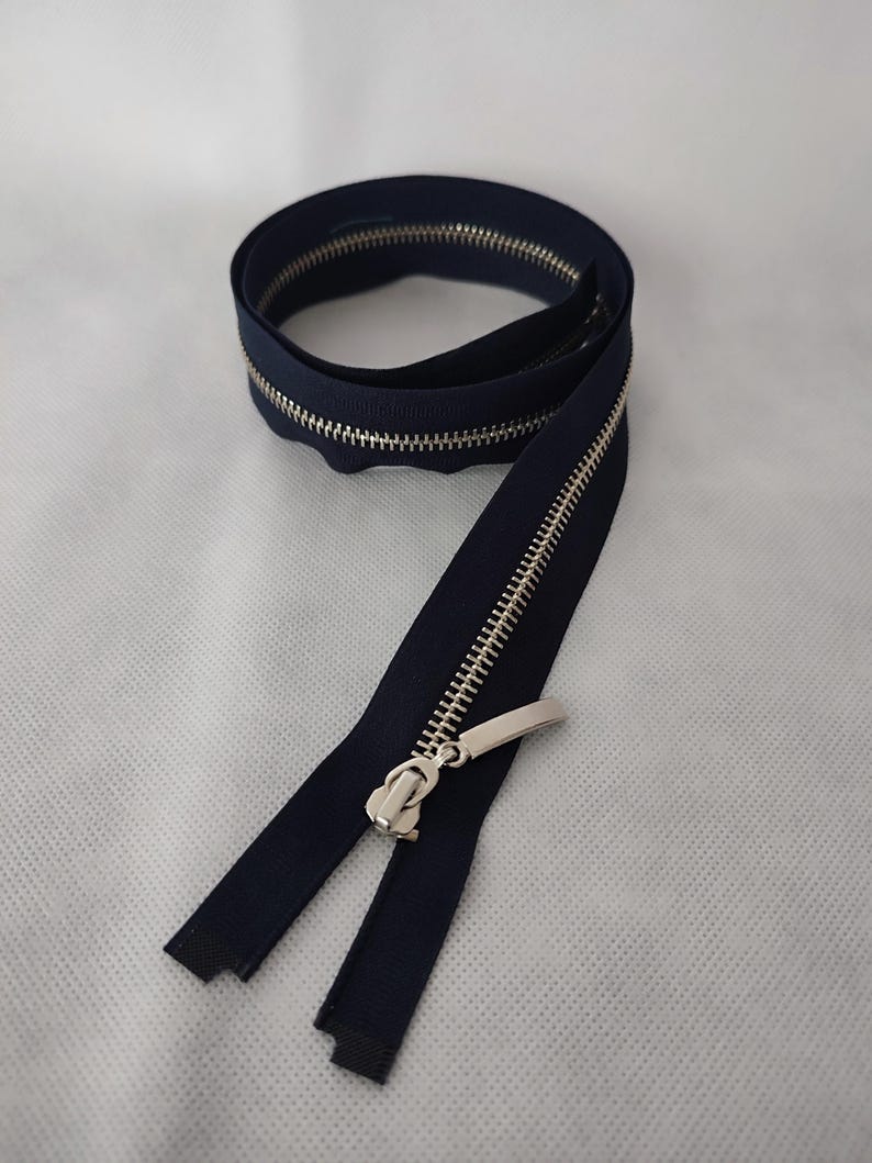 May include: A navy blue zipper with a silver metal pull and teeth. The zipper is coiled and partially unzipped, showing the metal teeth. Suitable for sewing projects and garment construction.