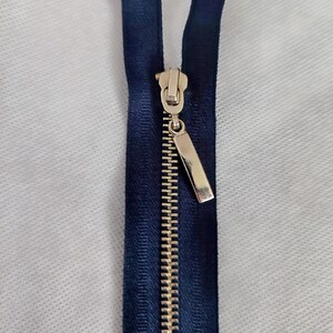 May include: A navy blue zip with a gold-coloured metal zip pull. The zip teeth are also gold-coloured. The zip is open, revealing the teeth and the fabric tape. The background is a white, textured surface.