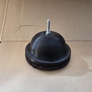 May include: A black, dome-shaped component with a threaded rod and nut protruding from the top. The base features a textured, dark brown surface. This is likely a part for machinery or equipment.