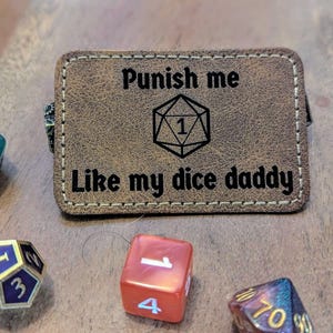 May include: A brown leather patch with the text "Punish me like my dice daddy" and a black and white illustration of a dice with the number 1 on it.