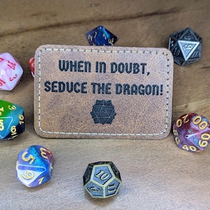 Könnte beinhalten: Ein brauner Lederkartenhalter mit dem Text "WHEN IN DOUBT, SEDUCE THE DRAGON!" Der Kartenhalter ist von bunten Würfeln umgeben.