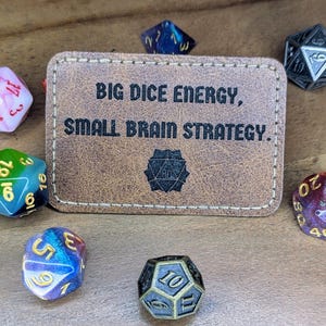 Può includere: Un porta carte in similpelle marrone con il testo "BIG DICE ENERGY, SMALL BRAIN STRATEGY." Il porta carte è circondato da sei dadi colorati.