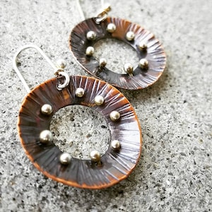 May include: A pair of copper earrings with a hammered texture and silver accents. The earrings are circular with a hole in the center and have a silver hook.