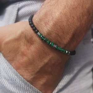 Op de afbeelding: Een kralenarmband met matzwarte kralen en levendige groene malachietkralen. De armband heeft ook zilverkleurige metalen accenten. De armband wordt om een pols gedragen, waarbij het ontwerp en het kleurcontrast worden getoond.