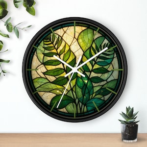 Puede incluir: Un reloj de pared redondo con marco negro. La esfera del reloj presenta un diseño de vidriera de hojas verdes y fondo amarillo. Las manecillas blancas indican la hora. Una pequeña planta en maceta está sobre una superficie de madera.