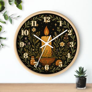 May include: A round wall clock with a light wood frame. The clock face is black with a candle illustration in the center, surrounded by floral and botanical elements. The clock hands are white.