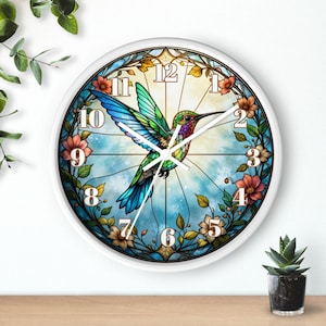 Puede incluir: Un reloj de pared con marco blanco que presenta un diseño de colibrí colorido. El colibrí está representado con tonos azules, verdes y morados, rodeado de detalles florales y un fondo azul claro. La esfera del reloj muestra números negros.
