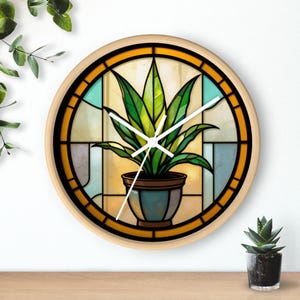 May include: A round wall clock with a stained-glass design featuring a potted plant. The clock face has a light wood frame, a yellow and blue background, and a green plant in a blue pot. The clock hands are white.