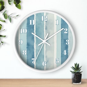 Pode incluir: Um relógio de parede redondo com moldura branca. O mostrador apresenta um design em aquarela de listras verticais em tons de azul e branco. Ponteiros e algarismos brancos.