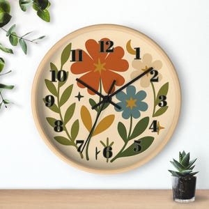 Puede incluir: Reloj de pared redondo con marco de madera clara. La esfera del reloj presenta un fondo crema con ilustraciones florales coloridas en naranja, azul y blanco. Las manecillas y los números negros completan el diseño, con una pequeña luna creciente en la parte superior.