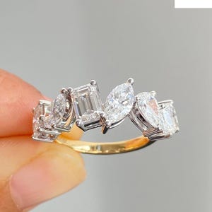 May include: A gold and diamond ring featuring a cluster of various-shaped diamonds. The ring has a gold band and multiple white diamonds in different cuts, including emerald, pear, and round shapes. The ring is held against a gray background.