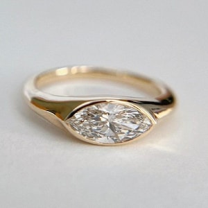 May include: A gold ring with a marquise-cut diamond. The ring has a smooth, polished band and a bezel setting that holds the diamond securely. The diamond sparkles brightly against a white background. The text "DEARTH JEWELS LONDON" is in the upper right corner.