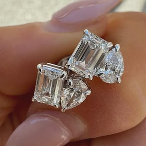 May include: Close-up of three diamond rings. The rings feature emerald, pear, and heart-shaped diamonds. The diamonds are set in a silver or white gold band. The rings are held up against a blurred background.