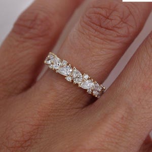 Könnte beinhalten: Ein goldener Ring mit einem Band aus abwechselnd tropfenförmigen und runden, klaren Edelsteinen. Der Ring wird an einem Finger getragen, mit dem Logo von Dearth Jewels London im Hintergrund.