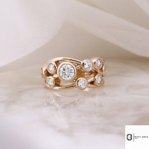 Könnte beinhalten: Ein goldener Ring mit mehreren klaren, runden Edelsteinen. Der Ring hat ein einzigartiges Design mit den Edelsteinen in einem fließenden, durchbrochenen Muster. Der Ring wird auf einer Marmoroberfläche präsentiert.