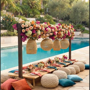 May include: A low wooden dining table set for an outdoor event, featuring woven rattan pendant lights suspended from a dark frame overhead. The table is decorated with floral arrangements, colourful napkins, and surrounded by textured floor cushions.