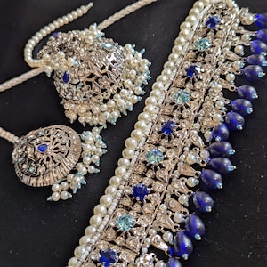 Puede incluir: Un conjunto de collar y pendientes plateados ornamentados. El collar presenta filas de perlas de imitación, piedras preciosas azules y transparentes, y cuentas azules colgantes en forma de lágrima. Los pendientes tienen un diseño a juego con perlas de imitación y piedras azules.