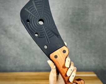 May include: A black cleaver with a wooden handle. The blade features a circular design with radial lines and three holes. The handle is made of light brown wood and has two dark brown dots.