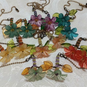 May include: A set of seven colorful floral necklaces with green leaves and beads. Each necklace features a different color flower, including pink, blue, green, orange, and yellow.