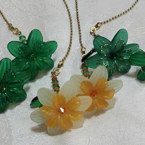 May include: Several flower-shaped pendant necklaces. The pendants are made of green and white translucent plastic with gold-colored chains. The green flowers have a darker green center, while the white flowers have an orange center.