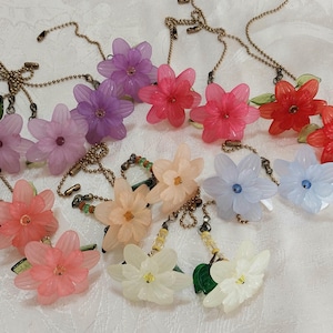 May include: A collection of floral necklaces in various colors, including pink, purple, red, and white. Each necklace features a translucent flower pendant with a delicate chain. The flowers have green leaf accents and are arranged on a white surface.