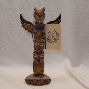 May include: A carved wooden totem pole featuring owl and human figures, painted in shades of brown, black, and red. The totem pole has a circular base and a paper tag with the text "Alaska Black Diamonds" attached with a blue ribbon.