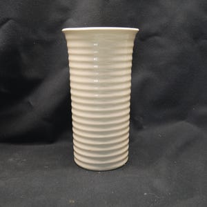 Puede incluir: Un jarrón cilíndrico de color crema con un diseño acanalado. El jarrón tiene un borde ligeramente ensanchado y una superficie lisa. El jarrón está hecho de cerámica o un material similar. Altura aproximada de 20 cm.