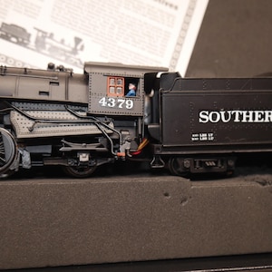 Puede incluir: Una locomotora de vapor a escala de color negro con el número 4379 impreso en el lateral de la cabina. El ténder lleva la inscripción SOUTHERN PACIFIC en letras blancas. Se ve una pequeña figura dentro de la ventana de la cabina.