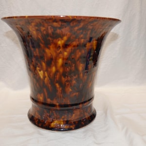 May include: A tall, flared vase with a glossy finish. The vase features a mottled pattern of brown, amber, and black, creating a tortoiseshell effect. The vase has a wide opening and a sturdy base, suitable for floral arrangements or decorative display.
