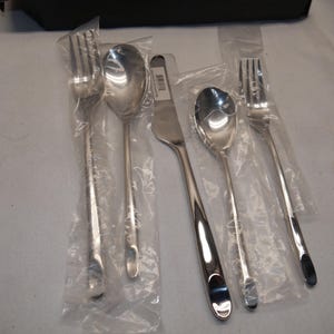 May include: A set of five stainless steel flatware pieces including two forks, two spoons, and one knife. Each piece features a smooth, polished finish with a rounded handle end. The items are individually wrapped in clear plastic protective sleeves.