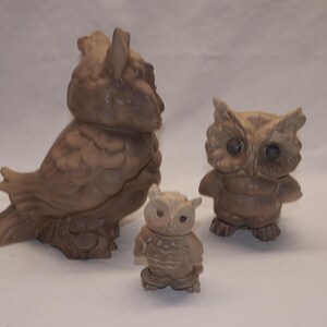 May include: A set of three brown owl figurines of varying sizes. The largest owl is positioned in profile facing left, while the medium owl faces forward with dark eyes. A smaller, third owl figurine stands in the center, also facing forward.