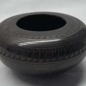 Könnte beinhalten: Eine runde, schwarze Keramikschale mit glatter, polierter Oberfläche. Das Äußere weist komplizierte, geätzte geometrische Muster auf, die aus sich wiederholenden horizontalen Linien und stilisierten Bogenformen bestehen, die den Körper umgeben.