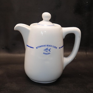 May include: White ceramic pitcher with a lid and handle. The pitcher has a blue stripe and the words "POMPANO BEACH CLUB BERMUDA" with a fish graphic. The pitcher is on a dark surface.