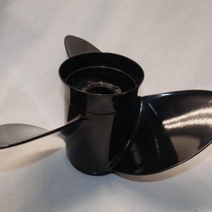 May include: A three-blade boat propeller with a glossy black finish. The central hub is cylindrical with a hollow core, featuring a visible internal gear mechanism. The blades are curved and smooth, extending outward from the central hub.